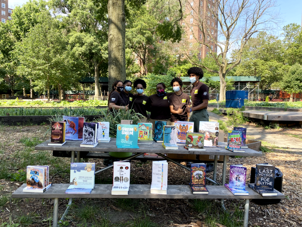 group of people standing outside behind a bench with books on it
