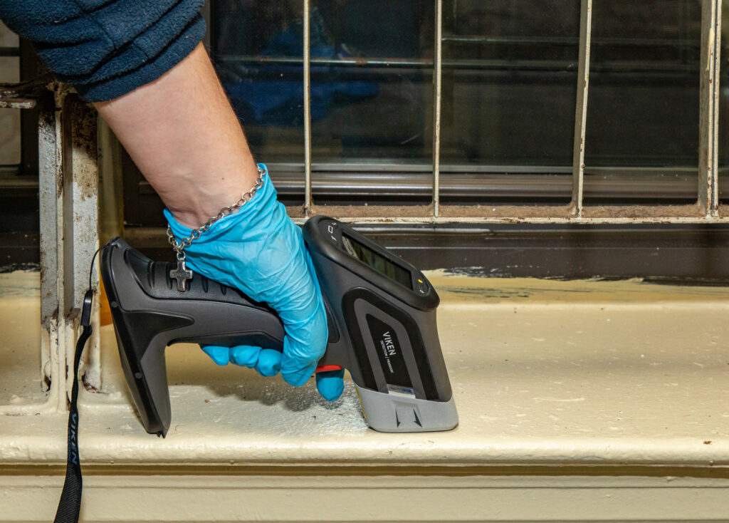 gloved hand holding an XRF analyzer (looks similar to a drill) to test paint on window sill
