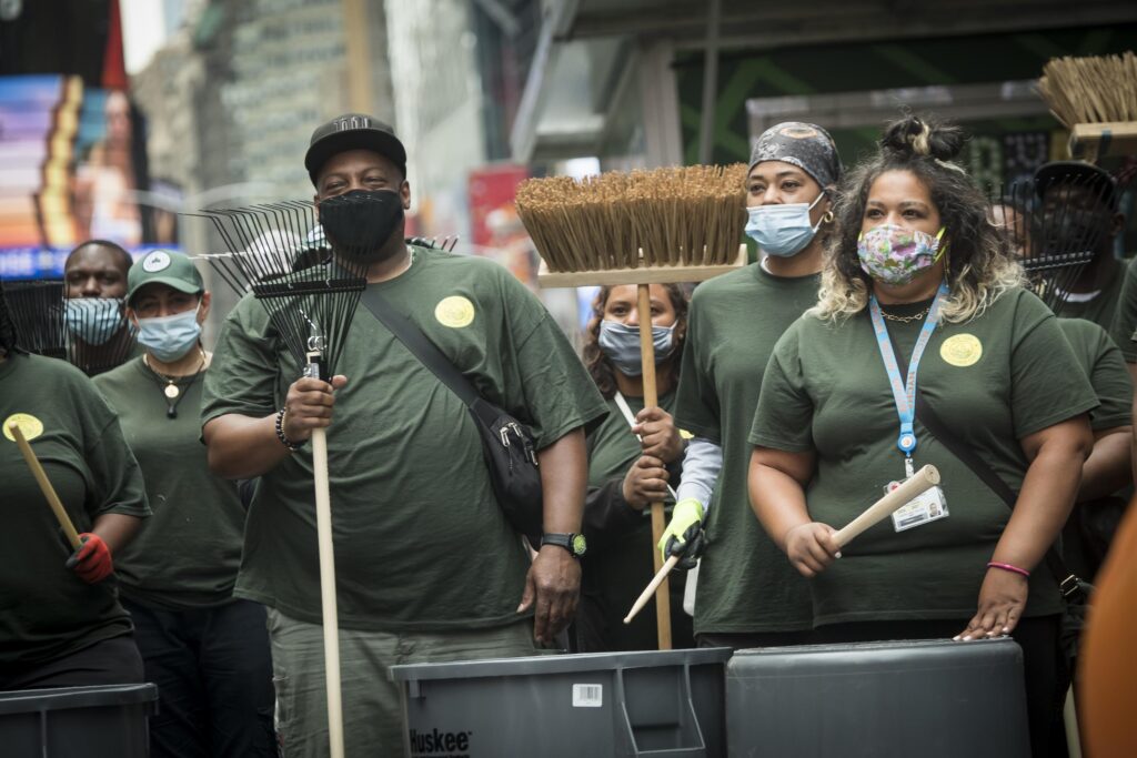 group of people in green shirts holding rakes, brooms, and trash cans.