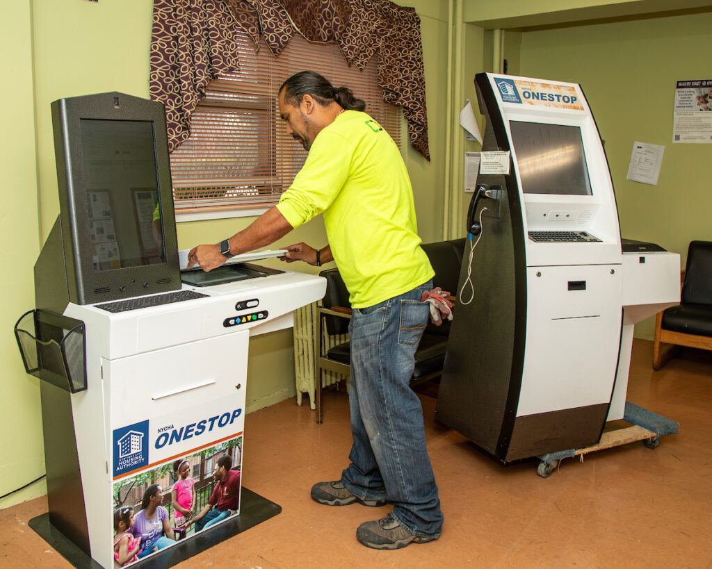 man installing self-service kiosk into a room. an older model of the kiosk is next to him