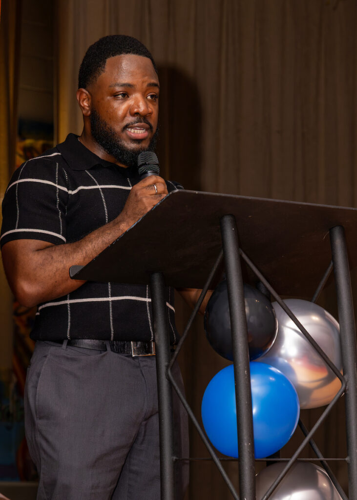 A person stands at a podium holding a microphone, speaking to an audience with balloons visible nearby.