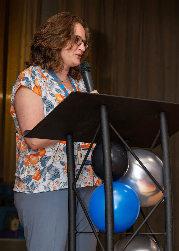 A person with curly hair and glasses stands at a podium, speaking into a microphone while wearing a floral shirt and holding a lanyard.
