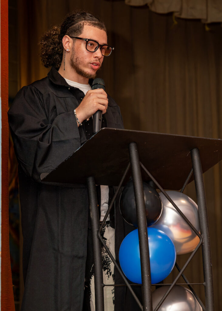 A person in a black robe stands at a podium with a microphone, speaking to an audience, while colorful balloons decorate the lectern.