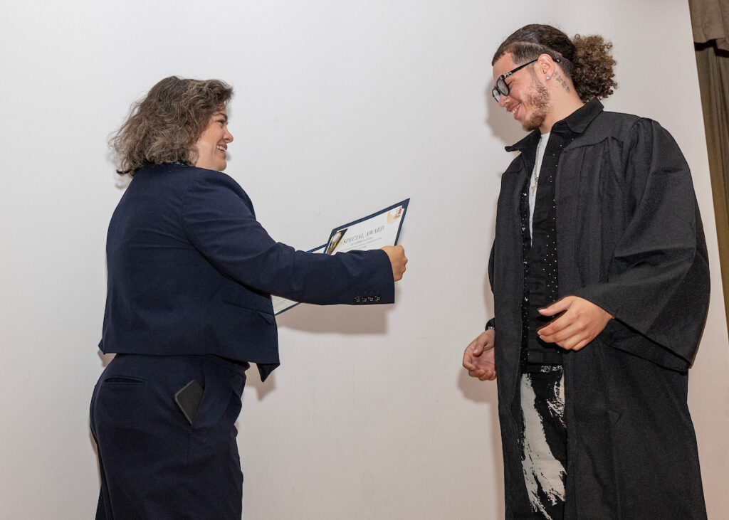 A person in a dark suit presents a special award certificate to another person dressed in a black robe, both smiling.
