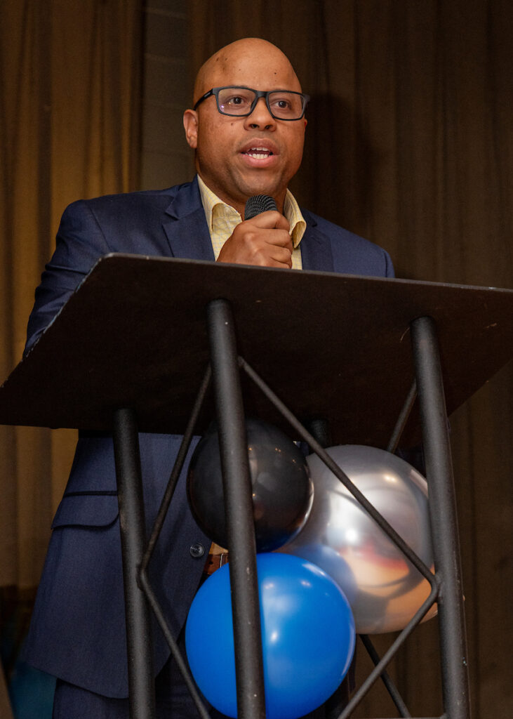 A person in a suit speaks into a microphone at a podium adorned with balloons.