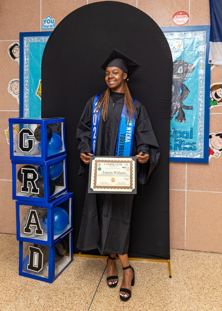 A young person in a graduation cap and gown stands proudly holding a certificate, with “GRAD” blocks and colorful decorations in the background.