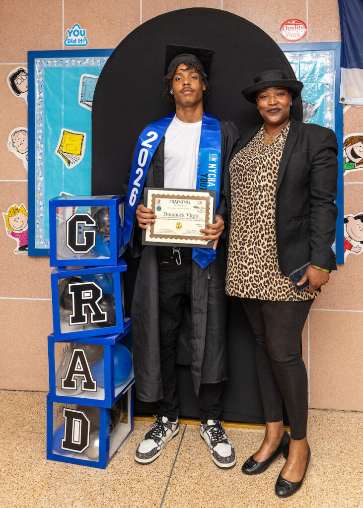 A young person in graduation attire holds a certificate and stands beside an adult next to a “GRAD” display, celebrating an academic milestone.