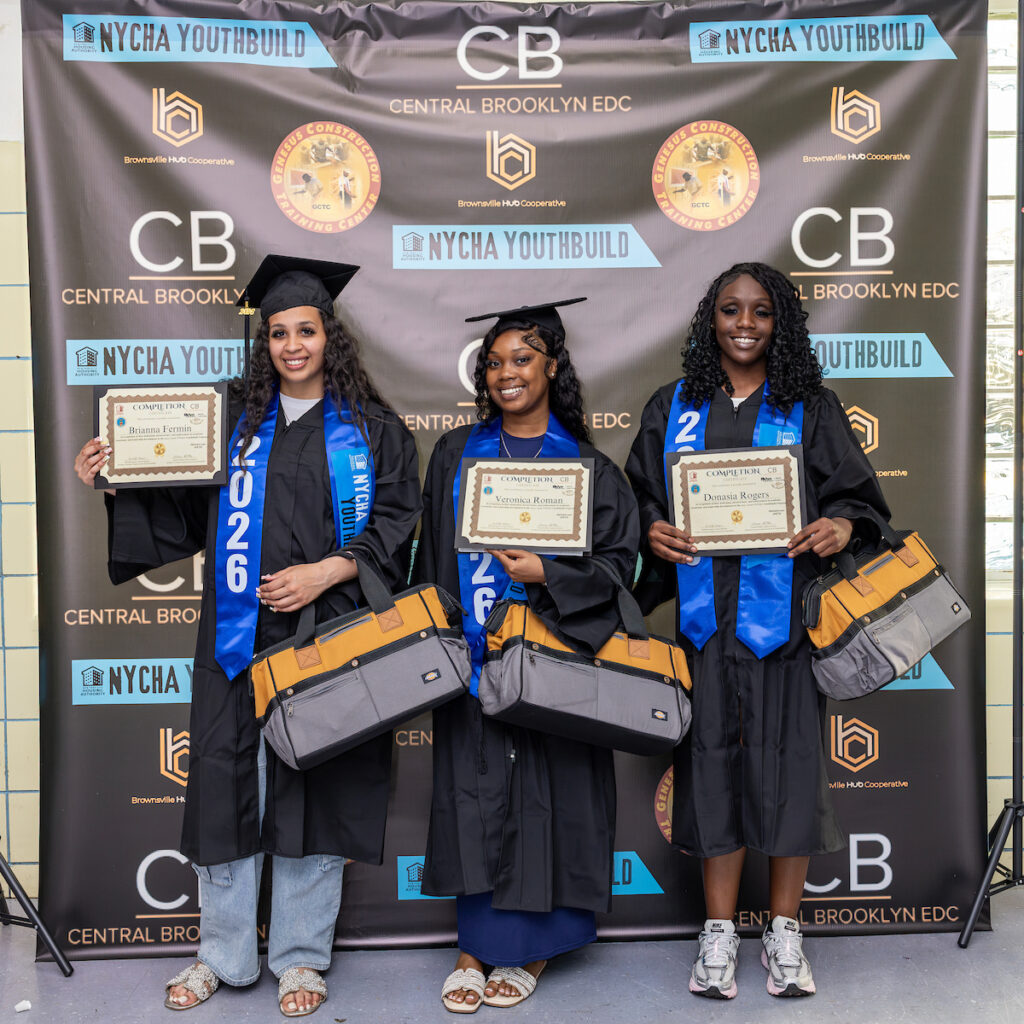 Three individuals in graduation attire stand before a backdrop featuring logos for NYCHA YouthBuild and Central Brooklyn EDC, each holding a certificate and smiling for the photo.