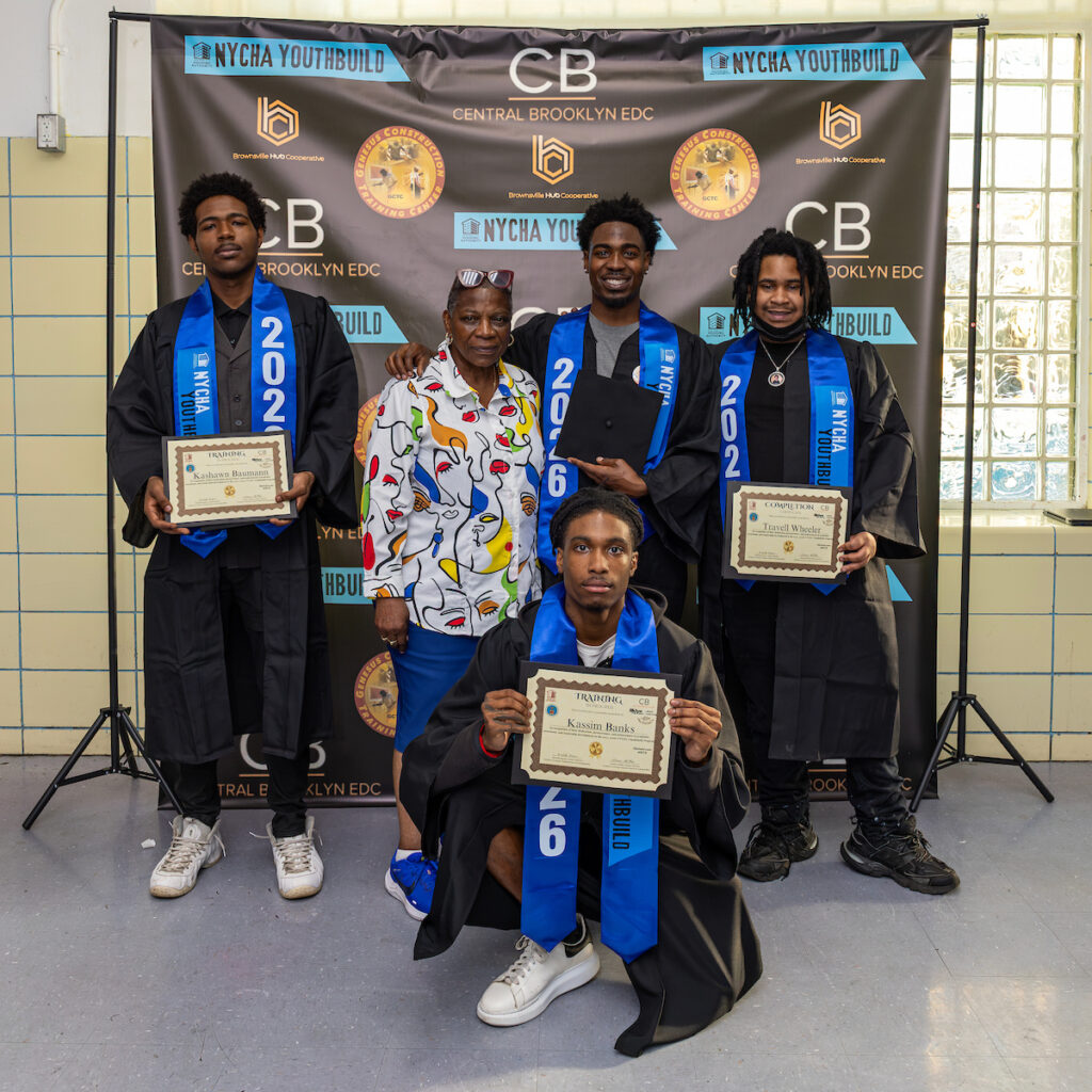 Four individuals in graduation attire and one person in a colorful shirt pose with certificates in front of a backdrop featuring NYCBA YouthBuild and Central Brooklyn EDC logos.