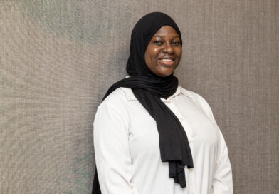 A person wearing a black hijab and a white shirt smiles warmly while standing against a textured gray wall.