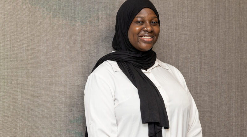 A person wearing a black hijab and a white shirt smiles warmly while standing against a textured gray wall.