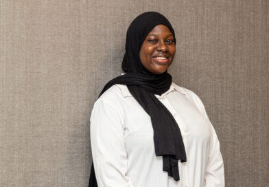 A smiling person wearing a black hijab and a white shirt stands against a textured gray wall.