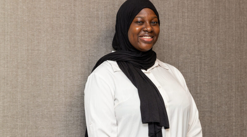 A smiling person wearing a black hijab and a white shirt stands against a textured gray wall.