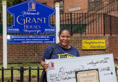 A person stands outdoors holding a large ceremonial check for $1,000 and an award certificate, in front of a NYCHA sign for Grant Houses.