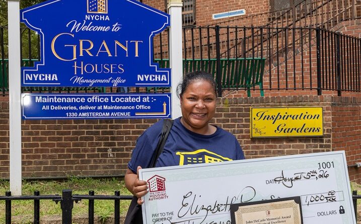 A person stands outdoors holding a large ceremonial check for $1,000 and an award certificate, in front of a NYCHA sign for Grant Houses.
