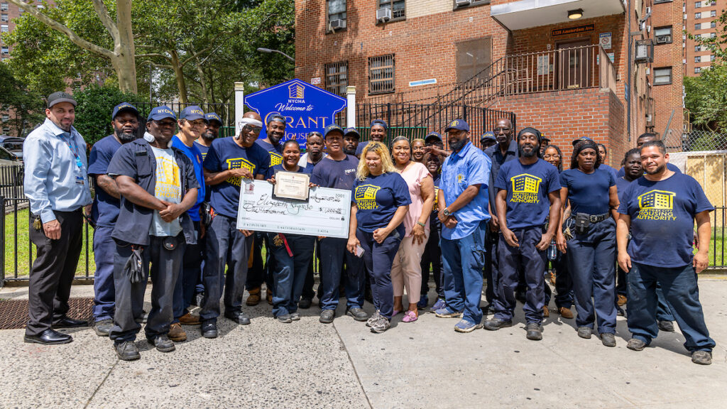A group of individuals, including some in uniforms and others in casual attire, pose outdoors holding a large ceremonial check and a plaque, standing in front of a brick building with a "Welcome to Grant" sign.