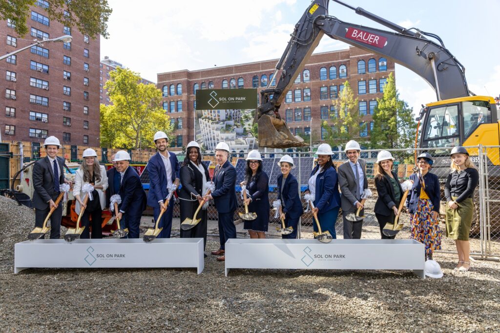 A group of individuals in hard hats stands at a construction site, holding shovels during a groundbreaking ceremony for "Sol on Park," with an excavator and brick buildings in the background.