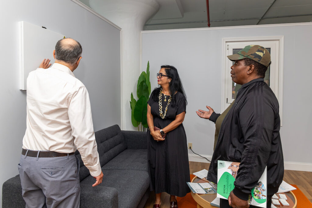 Three individuals engage in a conversation in a modern room, with one person gesturing while holding materials and another pointing at a whiteboard.