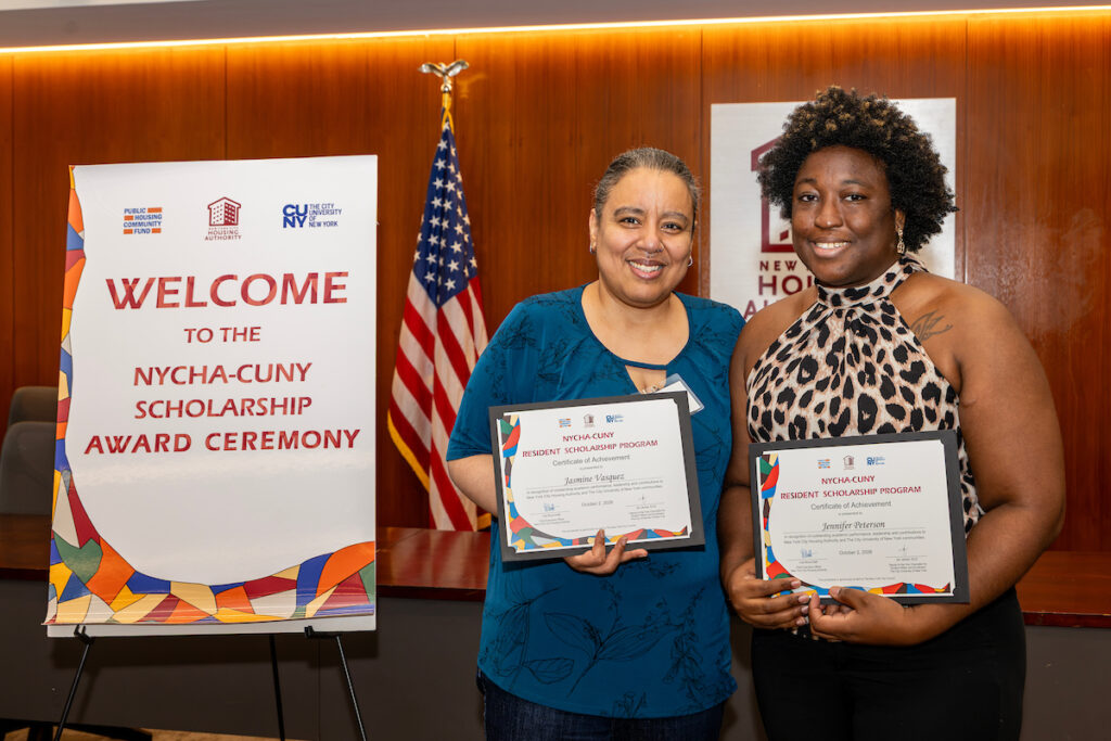 Two individuals stand together at an award ceremony, each holding a framed certificate from the NYCHA-CUNY Resident Scholarship Program, with a welcome banner and American flag in the background.