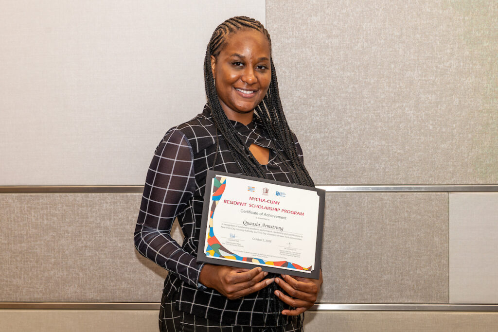 A person with braided hair smiles while holding a framed certificate from the NYCHA-CUNY Resident Scholarship Program.
