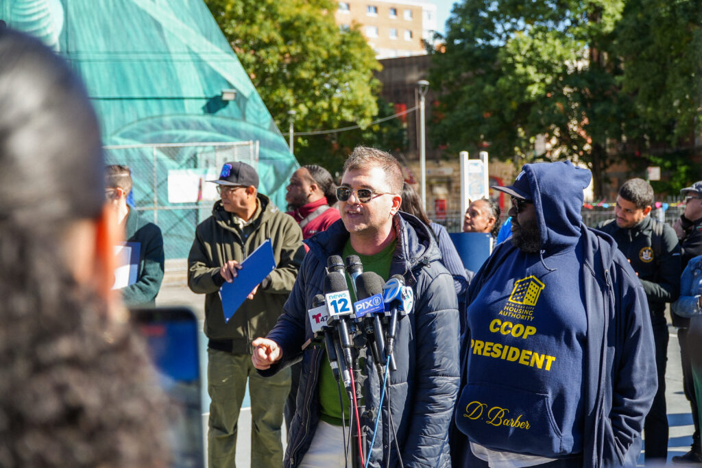 man in sunglasses talking into microphones, another man stands beside him