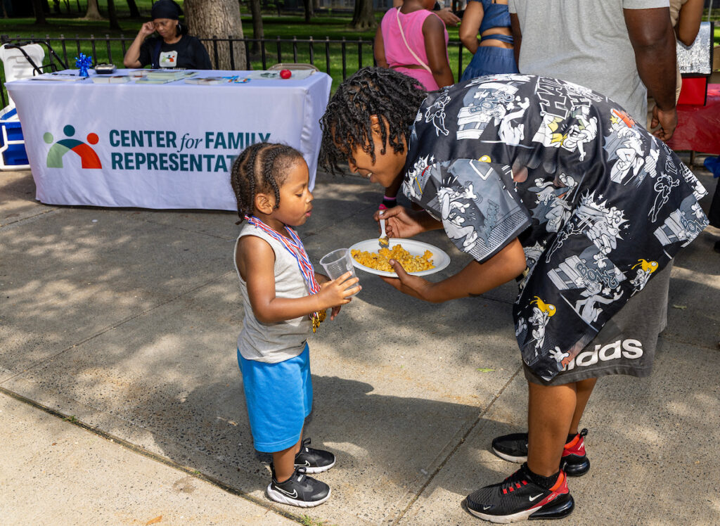 A young child stands on a sidewalk as an adult leans down to feed them food from a plate, with a table bearing the logo of the Center for Family Representation in the background.