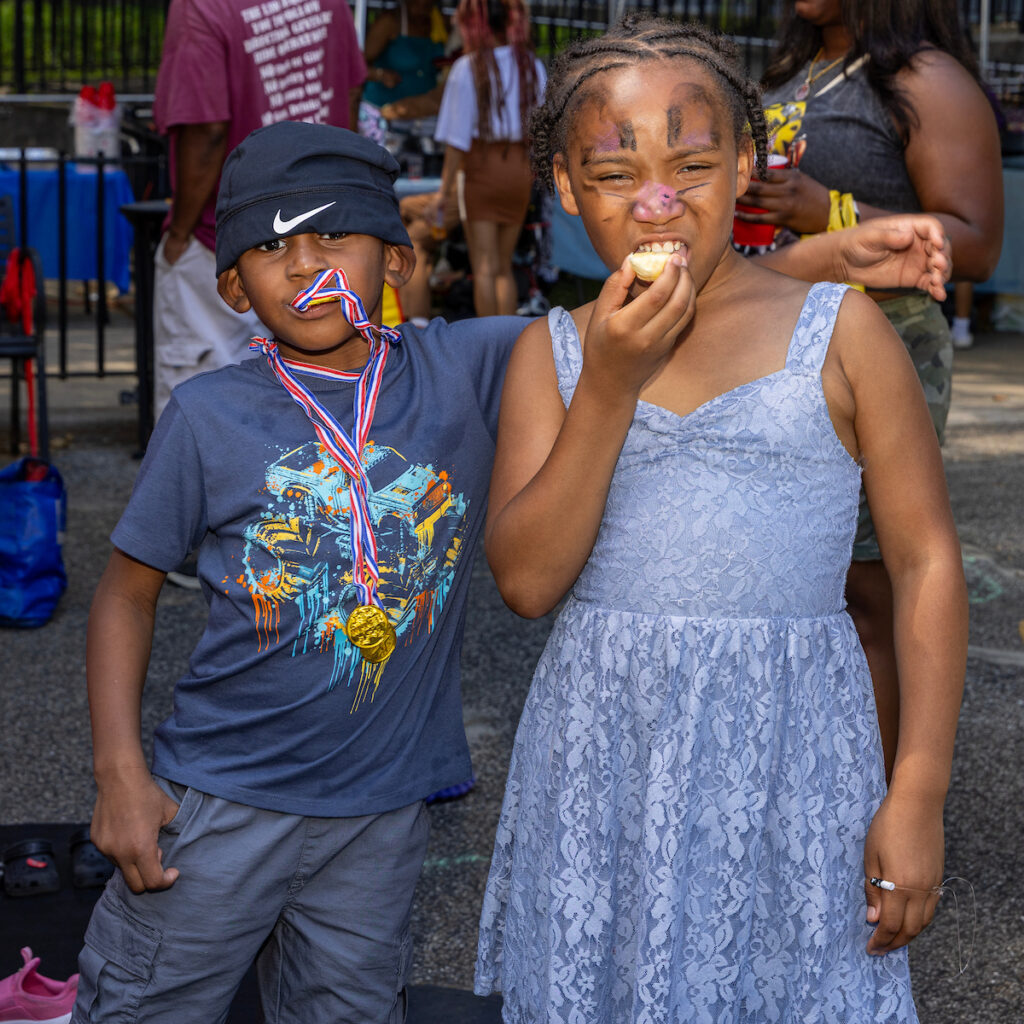 Two children stand together at an outdoor event, one wearing a medal and a hat with a ribbon, the other eating a snack while sporting face paint.
