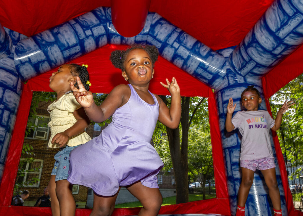 Three young children play joyfully inside a red and blue inflatable bounce house, with one making a peace sign and another blowing a bubble.