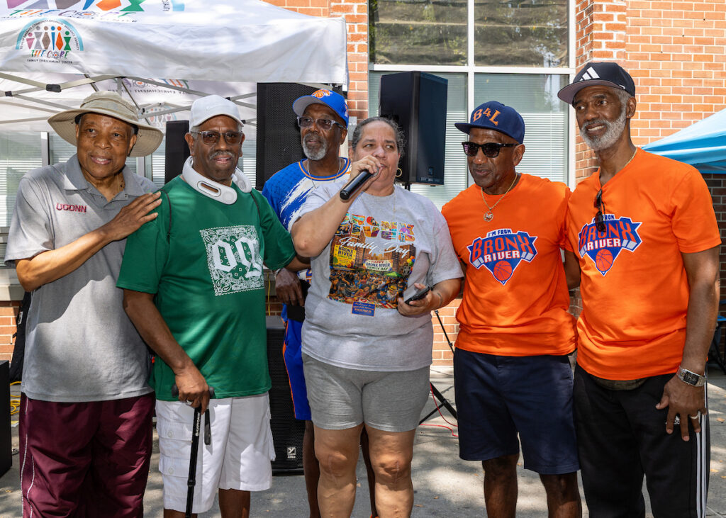A group of six people, including one holding a microphone, stand together outdoors at an event under a tent and brick building.
