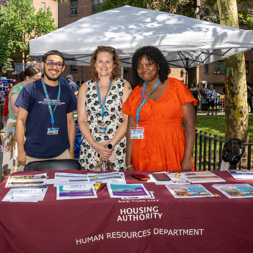 Three individuals stand behind a table at an outdoor event, promoting the New York City Housing Authority Human Resources Department.
