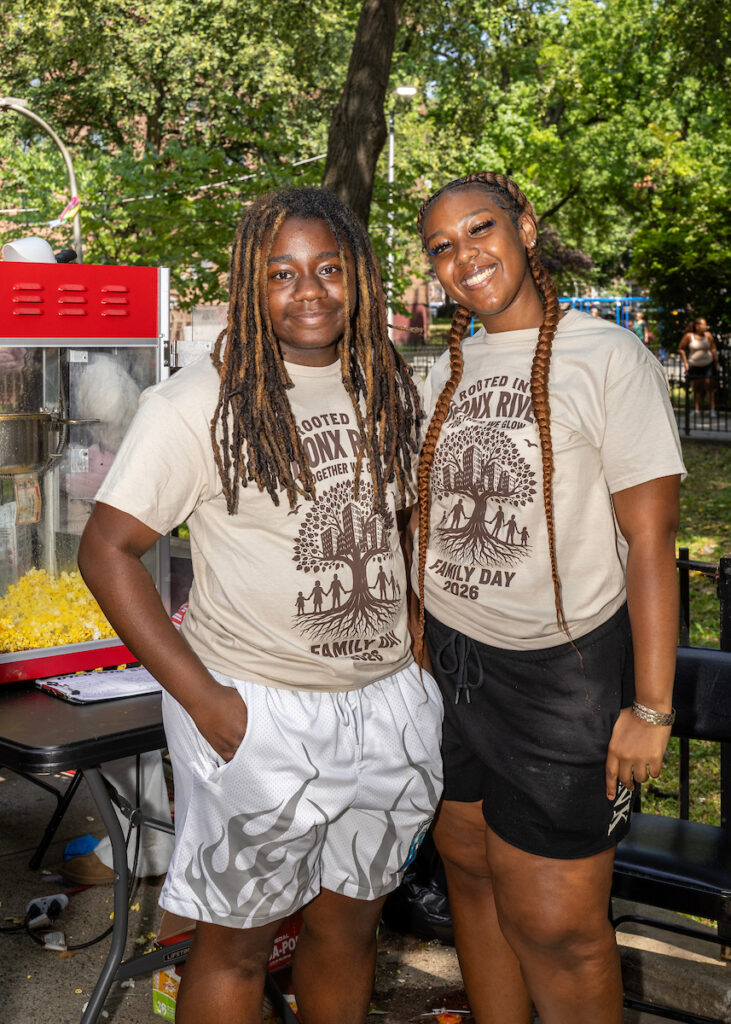 Two individuals wearing matching event shirts stand together outdoors near a popcorn machine, smiling for the camera.