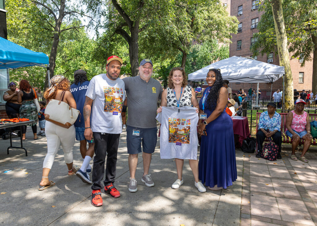 A group of four people stand together outdoors, one holding up a t-shirt with a colorful graphic, surrounded by trees and event tents with other attendees in the background.