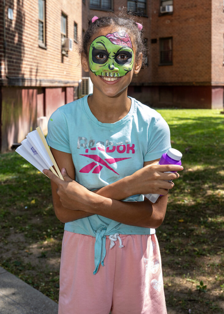 A child with vibrant green zombie face paint smiles while holding a fan and a purple bottle outdoors.