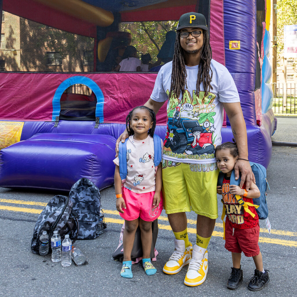A tall person with dreadlocks and a cap stands with two young children in front of a colorful inflatable bounce house, all smiling for the photo.