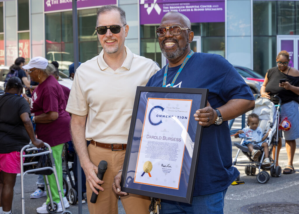 Two individuals stand outdoors, one holding a framed certificate, while others are visible in the background near a building with signage.
