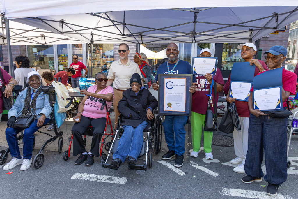 A group of people, including those using mobility aids, gather under a canopy to celebrate an event, with some holding framed certificates and awards.