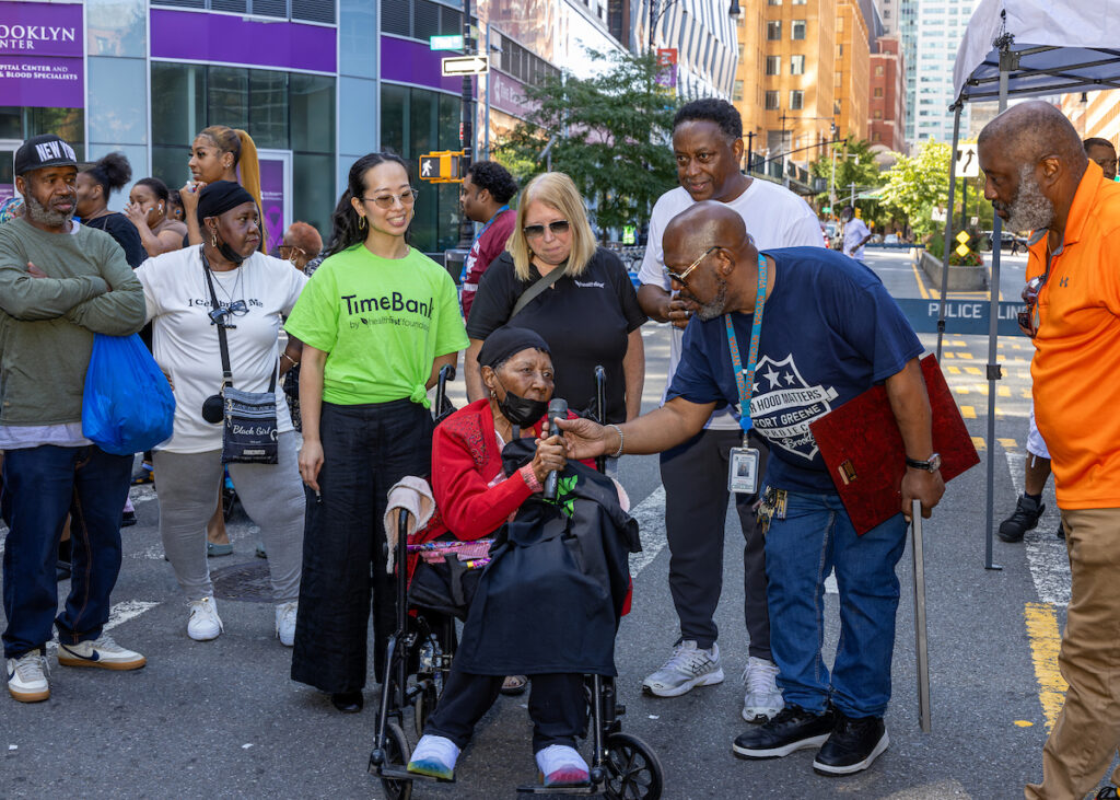 A group of people gathers around a person in a wheelchair on a city street, with one individual offering a microphone and another holding a cane, suggesting a public event or gathering.