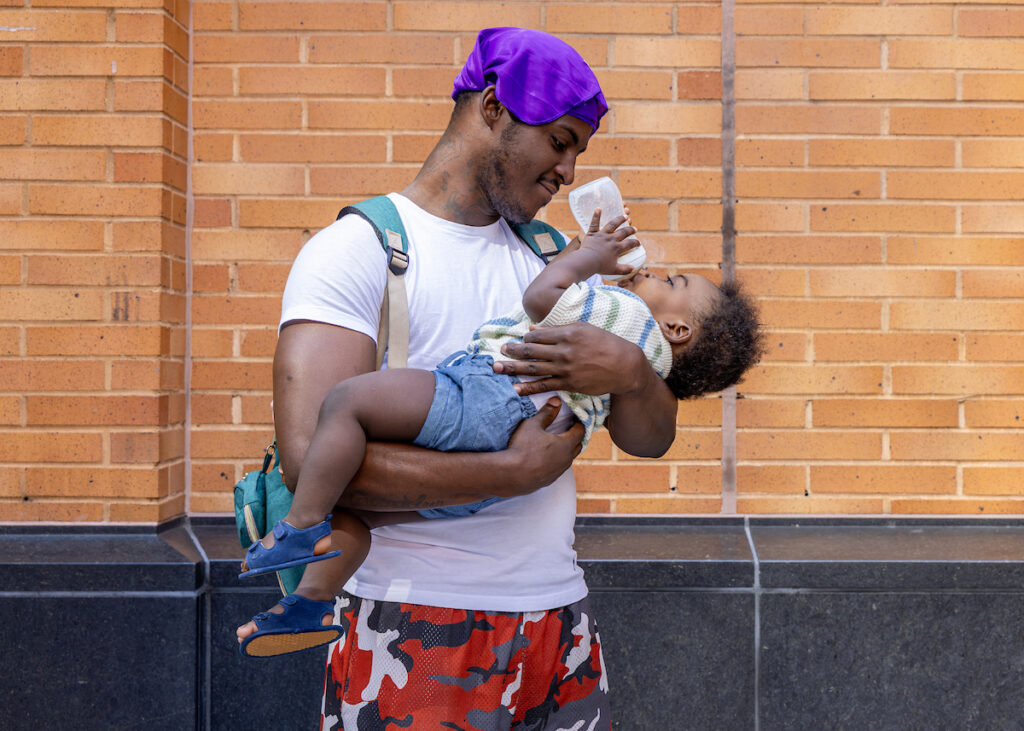 A person holds a baby while feeding them from a bottle, standing against a brick wall.