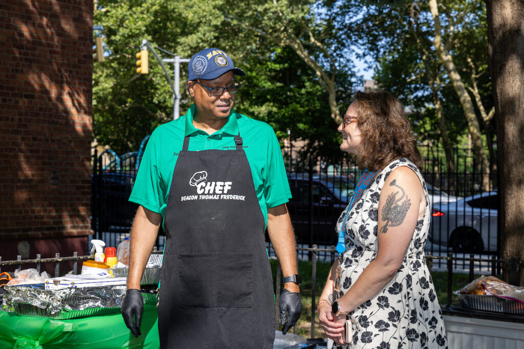 A chef in a green shirt and black apron stands outdoors beside a food station, conversing with a person in a floral dress who has a tattoo on their arm.