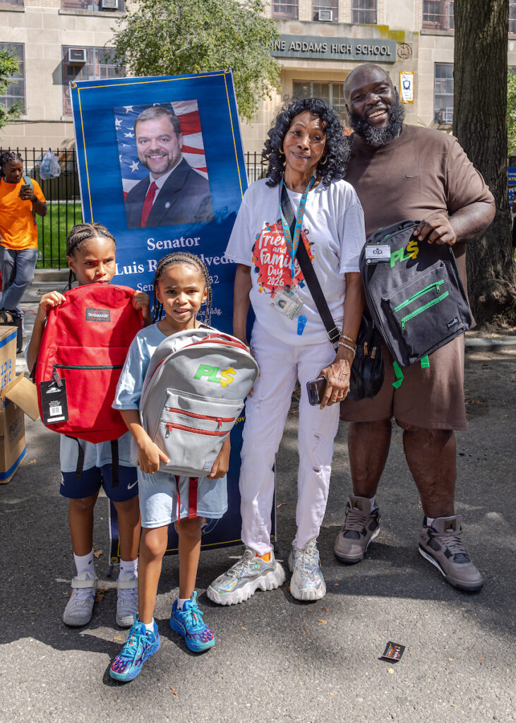 A family of four stands outdoors in front of a school, holding backpacks and posing beside a large poster of a political figure.