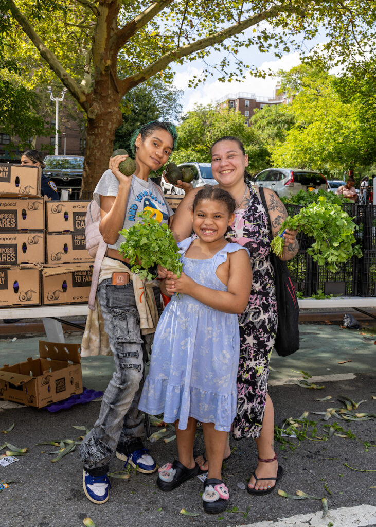 Three people stand together outdoors, holding fresh produce including avocados and cilantro, with stacked boxes of avocados behind them.