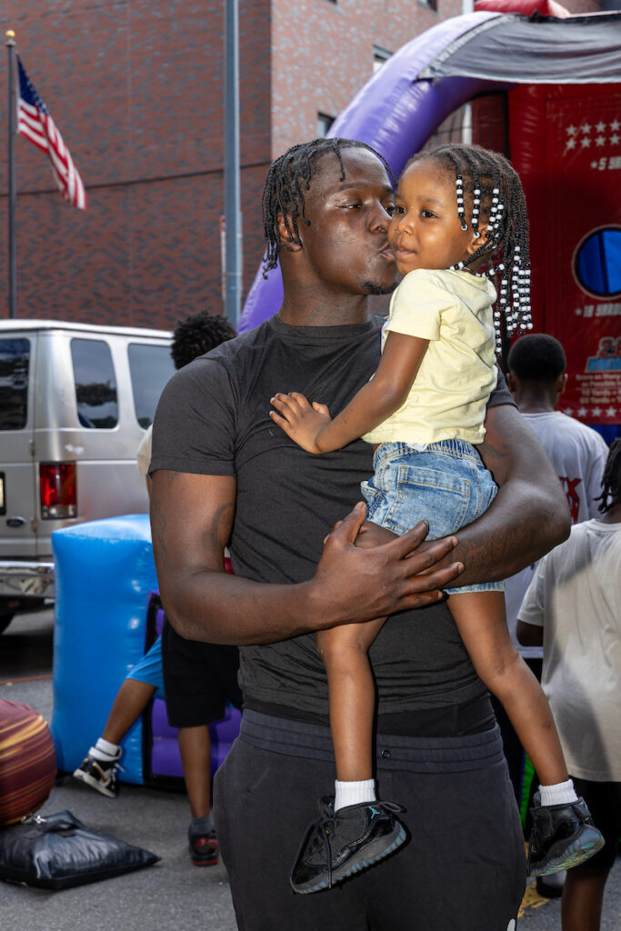 An adult holds a young child close while kissing their cheek at an outdoor event with inflatable structures and a flag in the background.