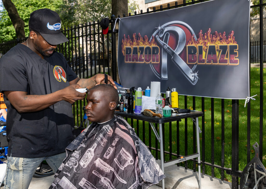 A barber styles a client's hair outdoors under a banner for "Razor Blaze" with grooming supplies on a table.