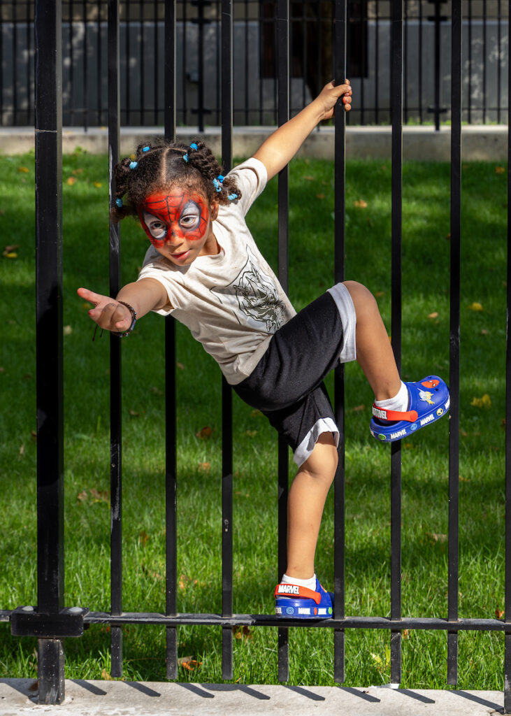 A child with Spider-Man face paint climbs a black metal fence, wearing Marvel-themed shoes and a superhero shirt.
