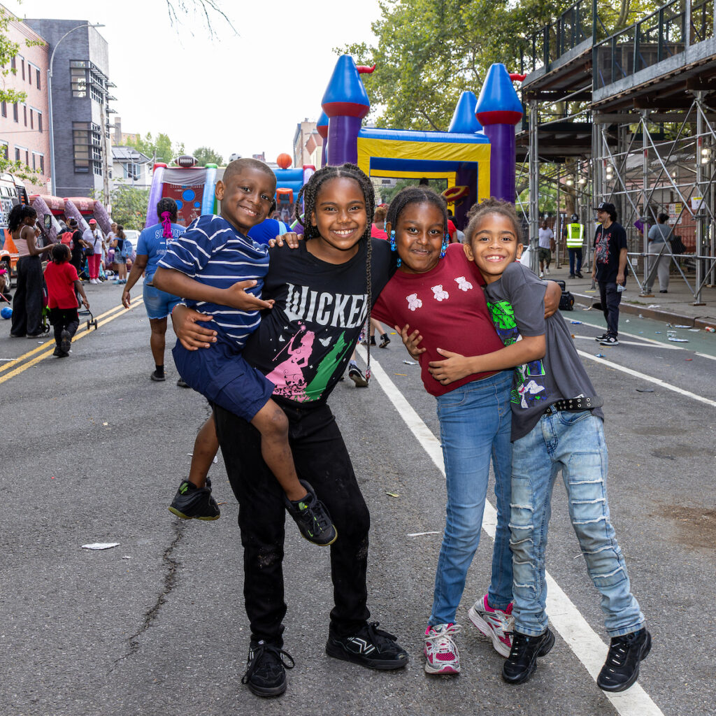 Four children smile for the camera while standing on a street during an outdoor event, with inflatable bounce houses and other people in the background.