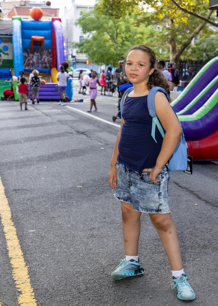 A young person stands on a street at an outdoor event, wearing a backpack and casual clothes, with inflatable play structures and other attendees visible in the background.
