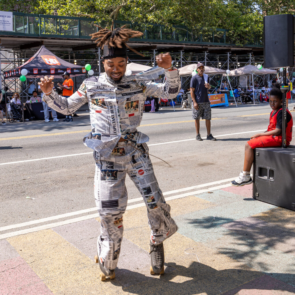 A person in a newspaper-themed outfit dances on roller skates in a street parade, with spectators and event tents in the background.