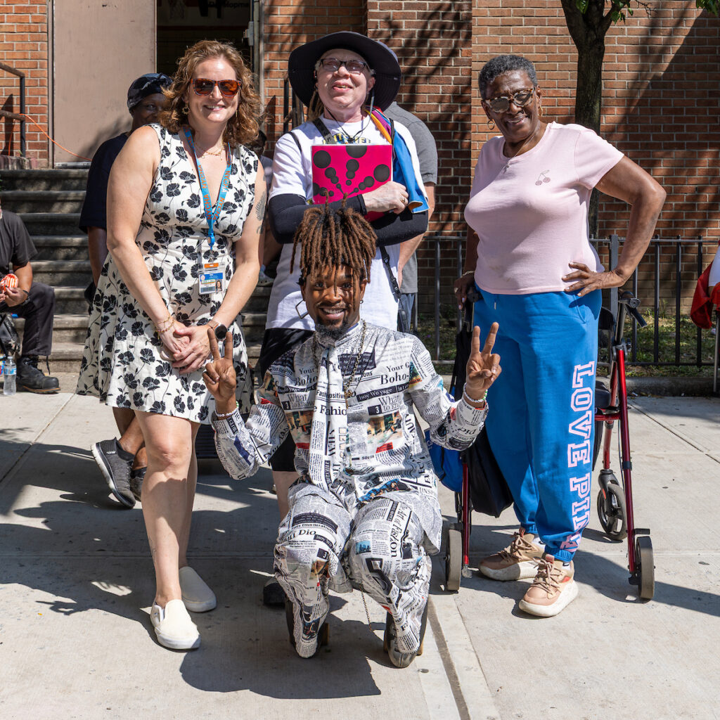 A group of five people pose outdoors on a sunny day, with one person kneeling in the foreground wearing a newspaper-print outfit and making peace signs.