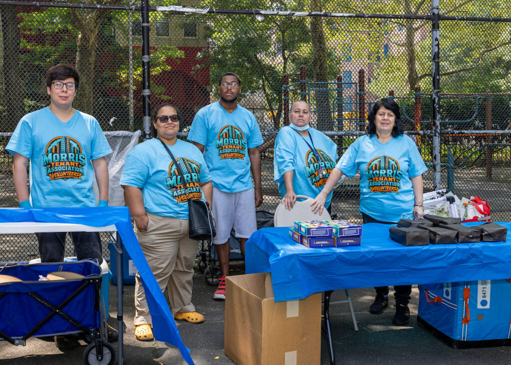 Five individuals wearing matching "Morris Tenant Association Volunteers" shirts stand behind tables with supplies at an outdoor event.