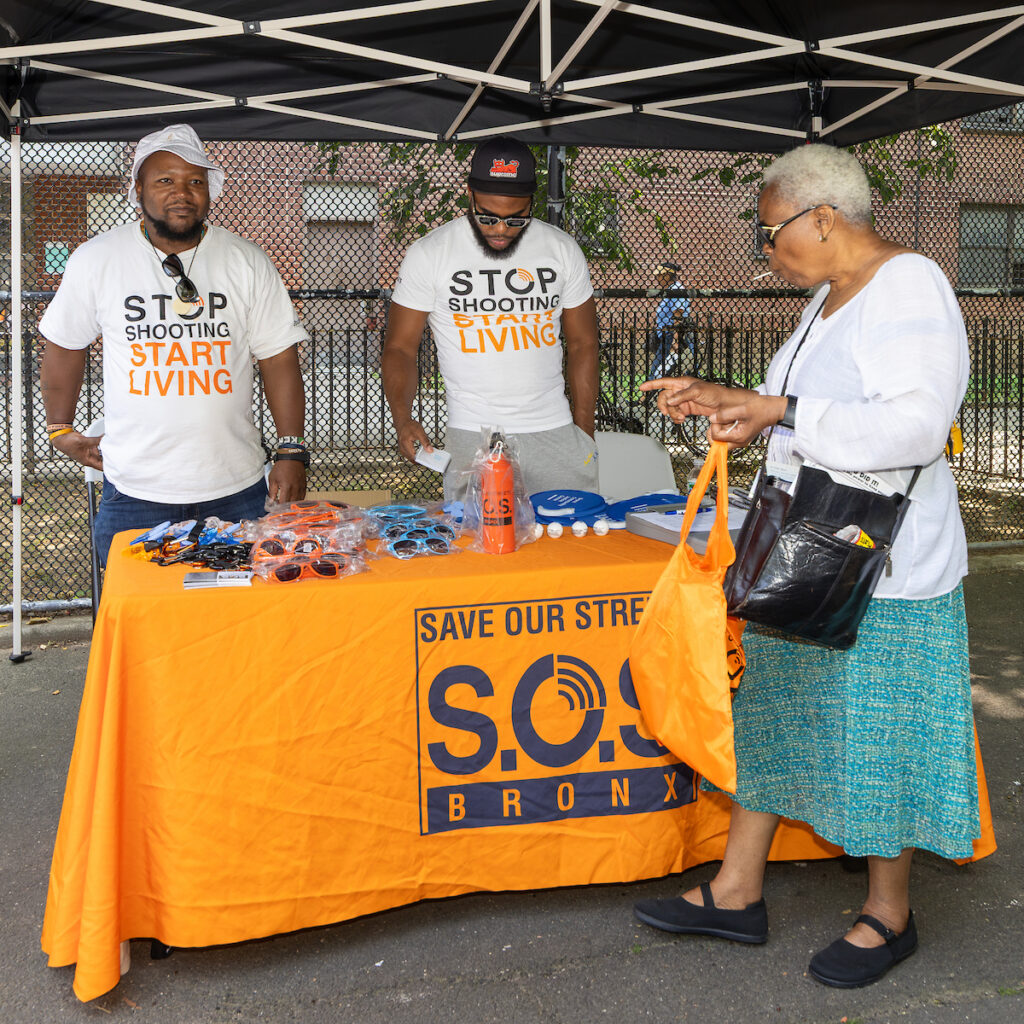 Two individuals in matching shirts stand behind a table with promotional items under a canopy, while a third person interacts with them, all part of a community outreach event for “Save Our Streets Bronx.”